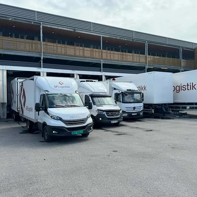 Flere hvite lastebiler med GF Logistikk-logo står parkert ved en lastehall.