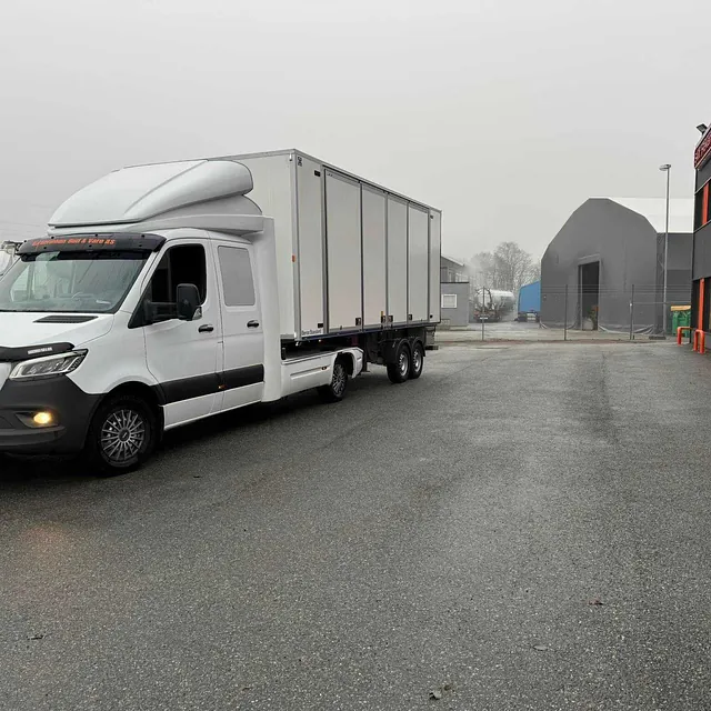 Hvit Mercedes lastebil med skaptilhenger parkert foran en industribygning i gråvær.