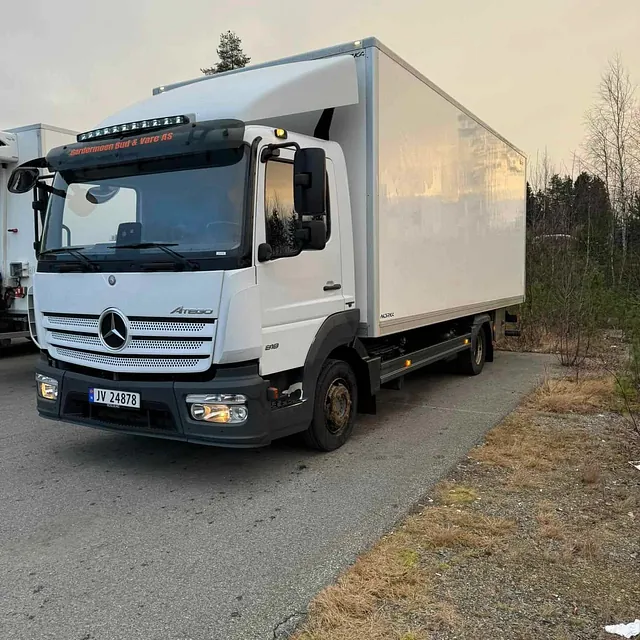 Stor hvit Mercedes Atego lastebil parkert på asfalt med skog i bakgrunnen.