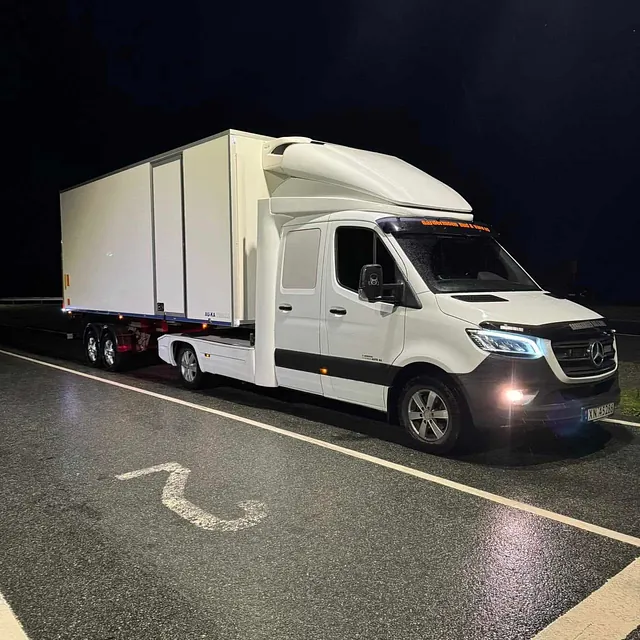 Stor hvit Mercedes lastebil med tilhenger parkert på en opplyst vei om natten.